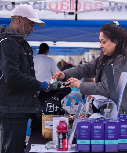 A woman places a bottle of hand sanitizer into a man's bag at a community event outdoors.